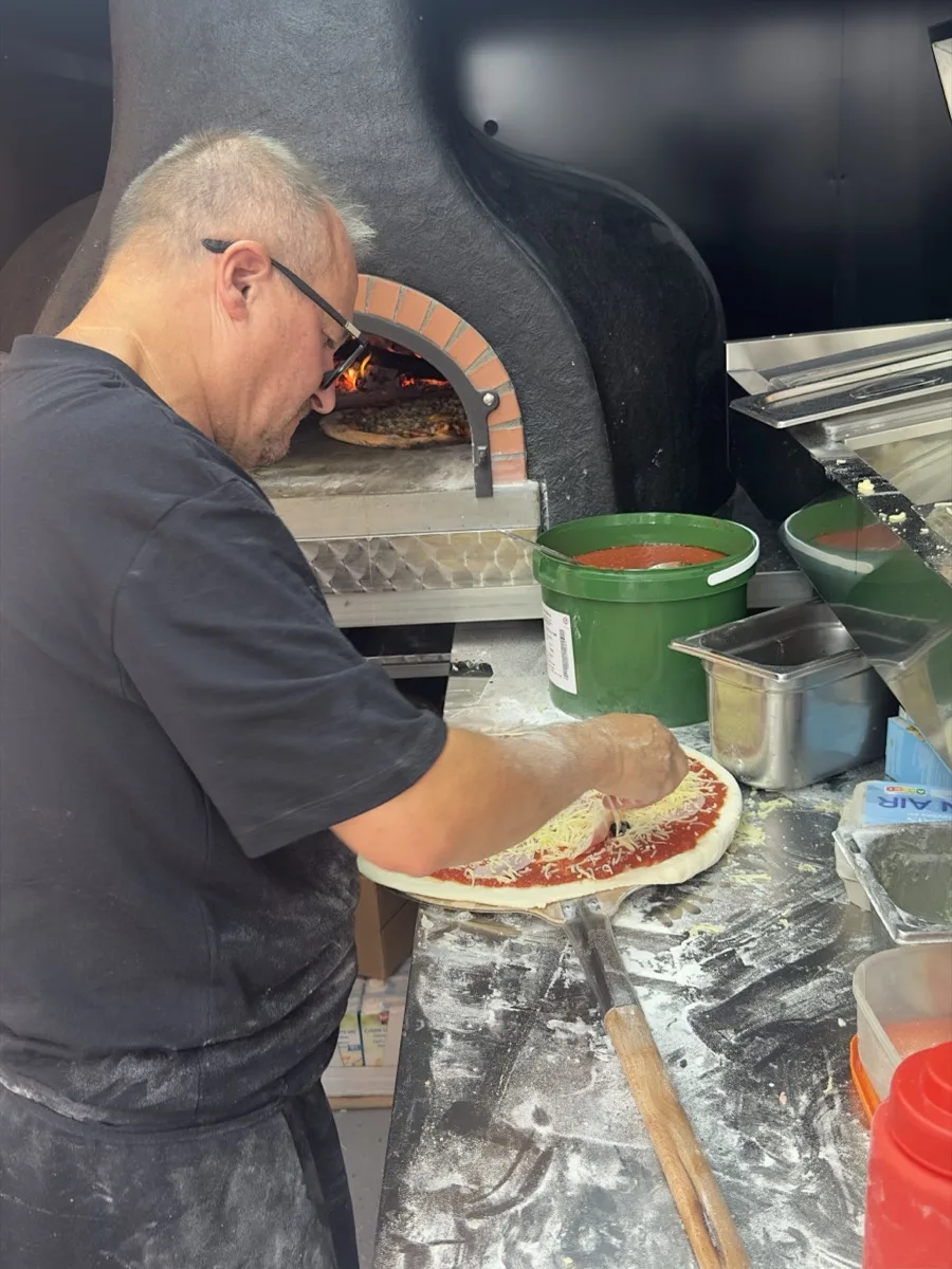 Tao garnit une pizza devant le four à bois allumé, dans le camion